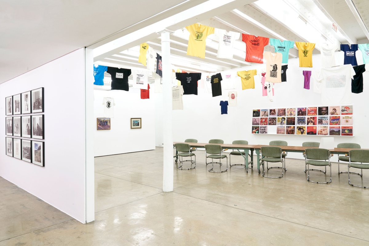 Installation photograph of the Common exhibition. At the back is a long wooden table. Above the table, T-shirts with slogans from the GALA Queer Archive and SAHA are suspended on criss-crossing lines. Behind the table, vinyl record covers from ‘The Library of Things We Forgot to Remember’ by Kudzanai Chiurai are arranged on a white shelf on the wall.
