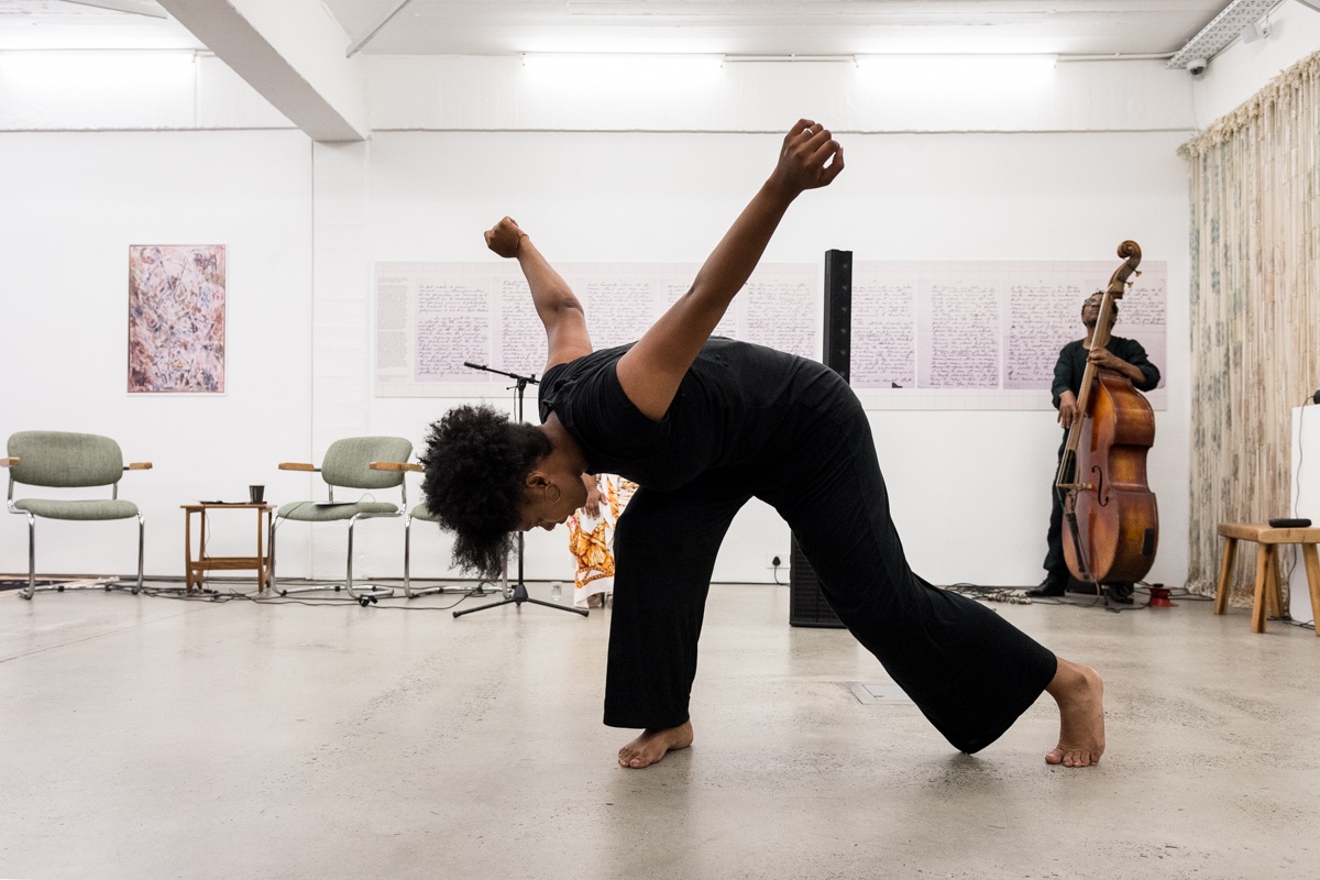 Event photograph from the Ernest Mancoba Symposium at A4 Arts Foundation. At the front, Amy Klaasen performs a dance. At the back, Herbie Tsoaeli plays the double bass.
