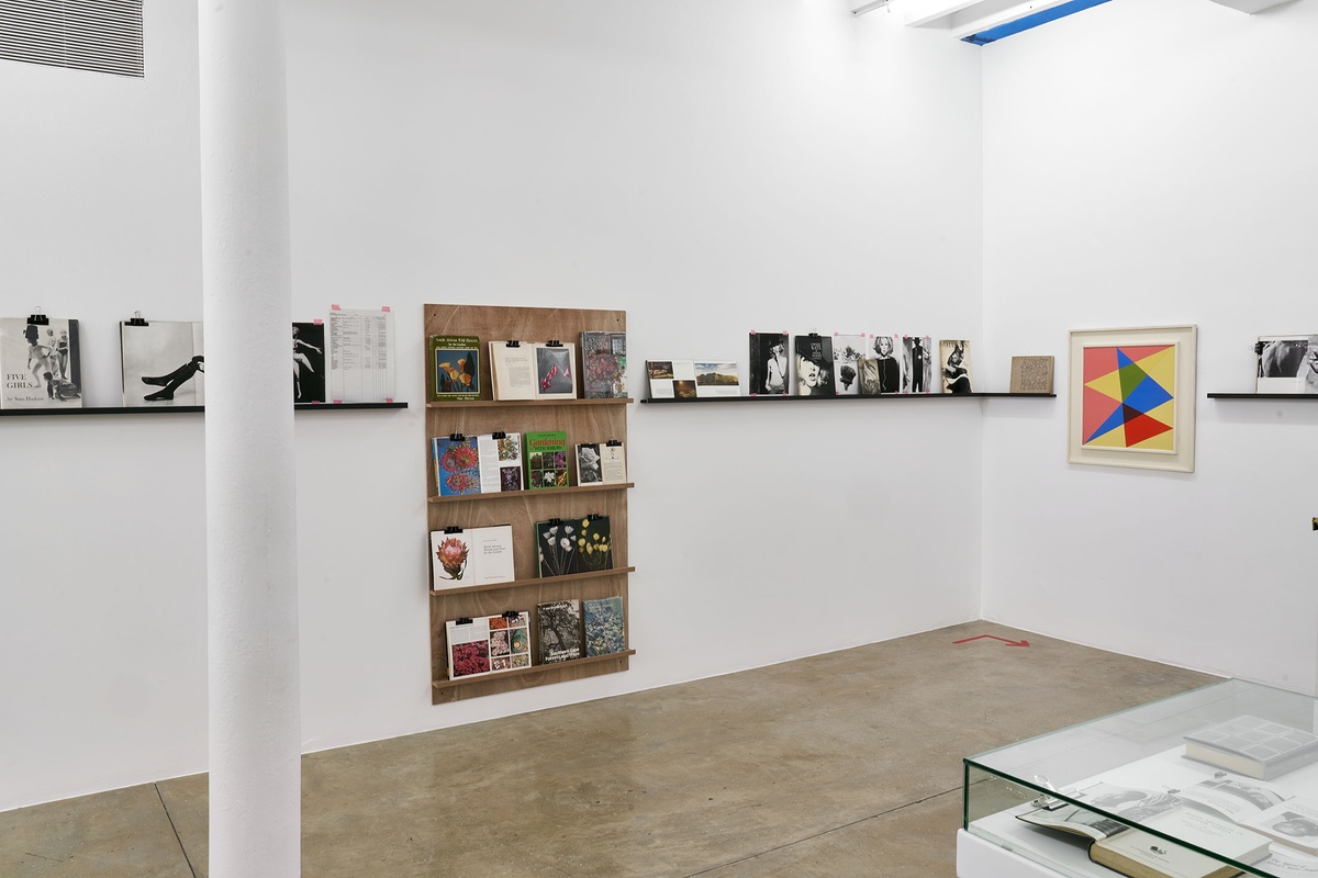 Installation photograph from the Photo Book! Photo-Book! Photobook! exhibition in A4’s Gallery. On the left, shelving units with printed matter from the years 1945 to 1967 line a white gallery wall. On the right, Albert Newall’s oil painting ‘Harmonic Development within a Square’ is mounted on a white wall.
