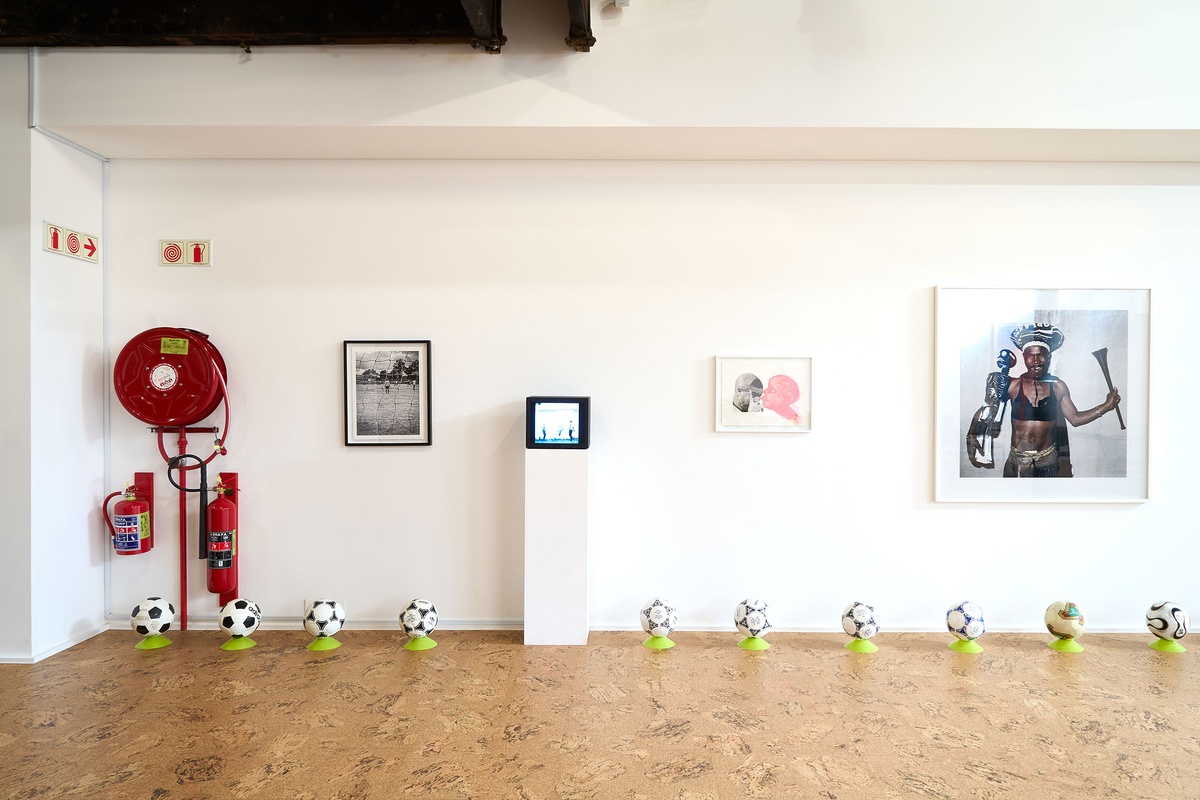 Installation photograph from the 2022 rendition of Exhibition Match on A4’s second floor. On the floor, soccer balls sit on plastic stands at the base of a white wall. On a white plinth, Robin Rhode’s animation ‘Hondtjie’ is displayed on a screen. On the wall are Andile Komanisi’s photograph ‘Soccer Indaba’, Penny Siopis’ lithograph ‘Pinky Pinky (Ronaldo)’ and Pieter Hugo’s photograph ‘Good Enough Mabaso, Orlando Pirates supporter, Coca-Cola Cup semi-final, Rustenburg, 2005’.
