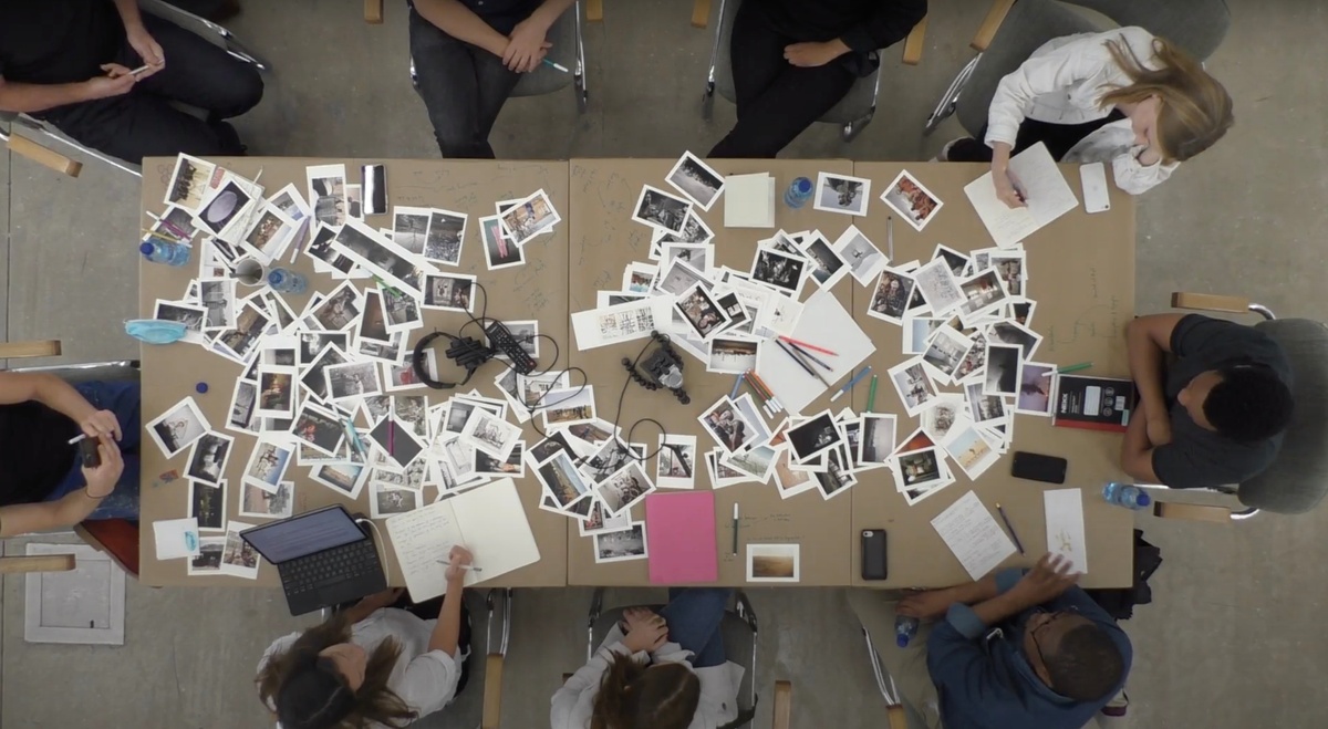 A still frame from the video recording of a roundtable discussion that accompanied the ‘Tell It to the Mountains’ exhibition in A4’s gallery. A top-down view of a table that is strewn with postcard-size reproductions of photographs by Lindokuhle Sobekwa and Mikhael Subotzky, with participants seated around it.
