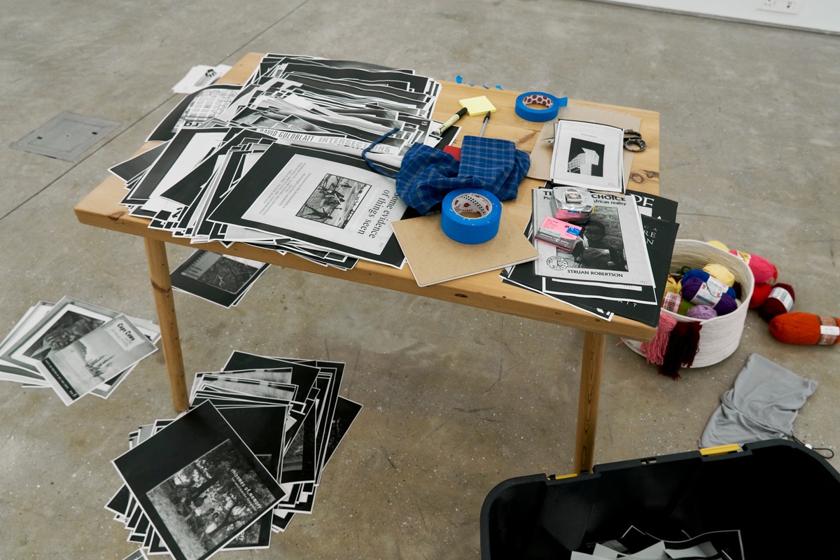 Process photograph from ‘Photobooks’, Sean O’Toole’s Course of Enquiry at A4. A wooden table is laden with photocopies from O’Toole’s photobook collection and rolls of blue tape. On the floor, bunches of variously coloured wool sits in a basket.
