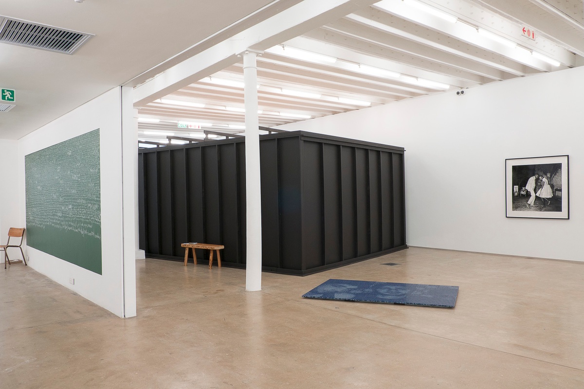 Installation photograph from the You & I exhibition in A4’s Gallery. On the left, a chalkboard with chalk writing from Brett Seiler and Luvuyo Nyawose’s performance installation ‘Reading Homophobia’ is mounted on the wall, with a single chair on the left. In the middle, Pierre Fouché’s indigo dice sculpture ‘Mahmoud & Ayaz’ sits on the gallery floor. On the right, Malick Sidibe’s monochrome photograph ‘Nuit de Noël (Happy Club)’ is mounted on the gallery wall.
