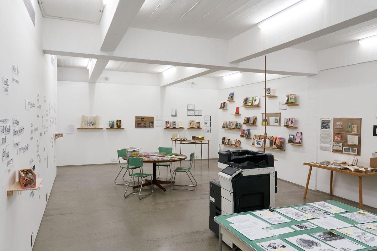 Installation photograph from the Papertrails exhibition in A4’s Reading Room. In the middle, a photocopier and round table. On the left, printed phrases are pasted onto the wall. At the back and on the right, the walls feature various shelves lined with printed matter.
