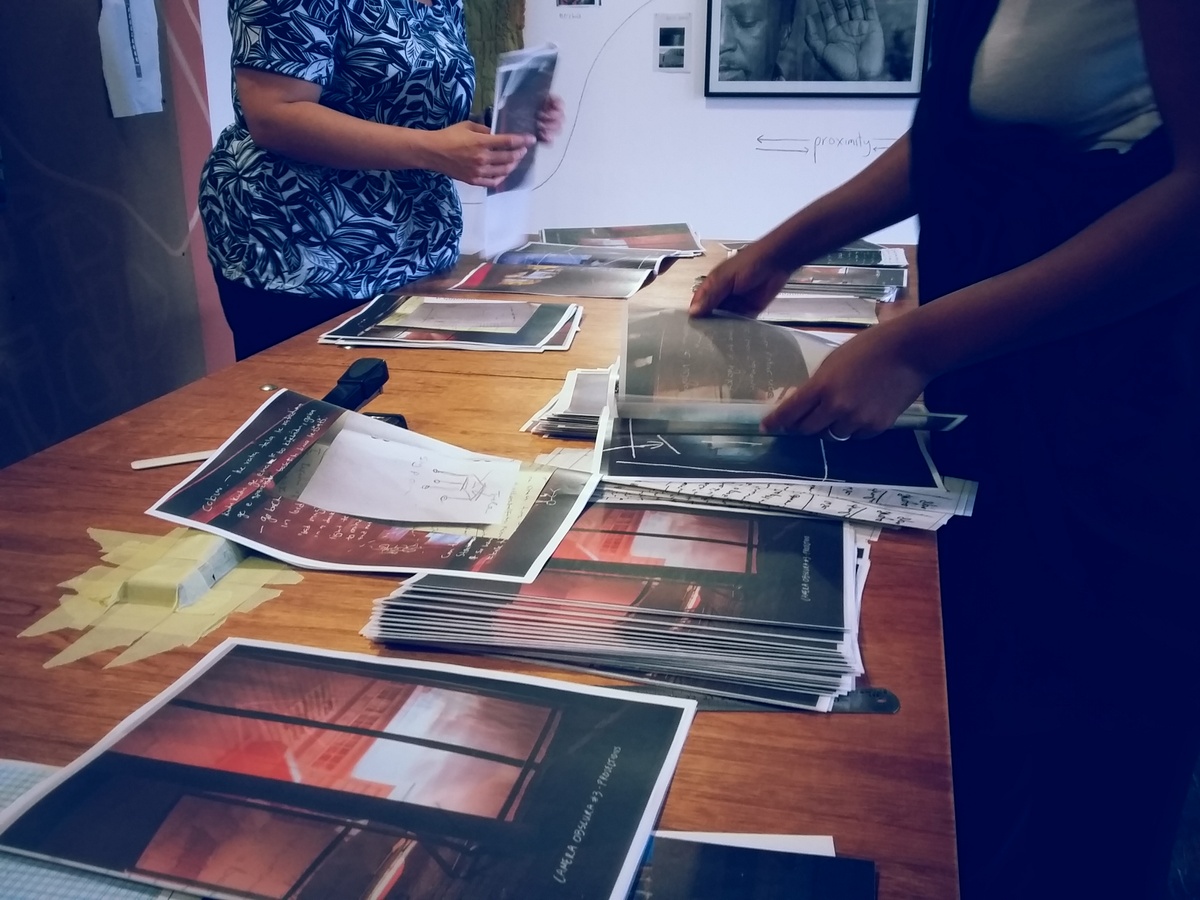 Ephemera from the making of francis burger’s publication for Tebogo George Mahashe’s ‘Camera Obscura #3’ exhibition on A4’s top floor that shows two individuals assembling the publication on a long wooden table. At the back, George Hallet’s monochrome photographic diptych ‘Peter Clarke’s Tongue’ is mounted on a white wall.
