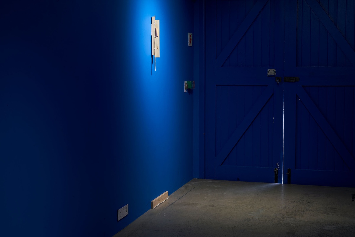 Installation photograph from Guy Simpson's Passage exhibition in A4 Arts Foundation's Goods space shows two sculptures that recall the mezuzah attached to the wall. At the front, one wood sculpture is painted blue to match the wall. At the back, another wood and electrical wire sculpture is unpainted.
