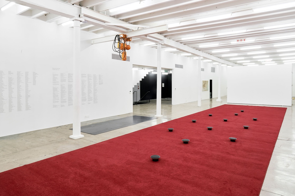 Installation photograph from The Future Is Behind Us exhibition in A4’s Gallery. In the middle, James Webb’s sound installation ‘Prayer’ features an long red carpet with upturned audio speakers laid out in a geometric pattern.
