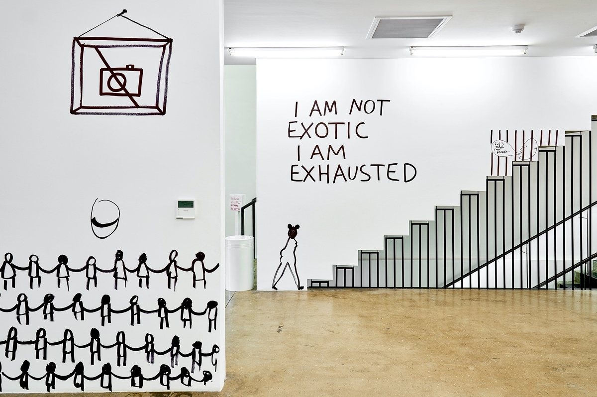 Installation photograph from Dan Perjovschi’s ‘The Black and White Cape Town Report’ exhibition in A4’s Gallery that shows black felt pen marker drawings on white walls. On the left, a drawing of a framed picture of a camera with a strikethrough line. At the back, the phrase ‘I am not exotic, I am exhausted’.
