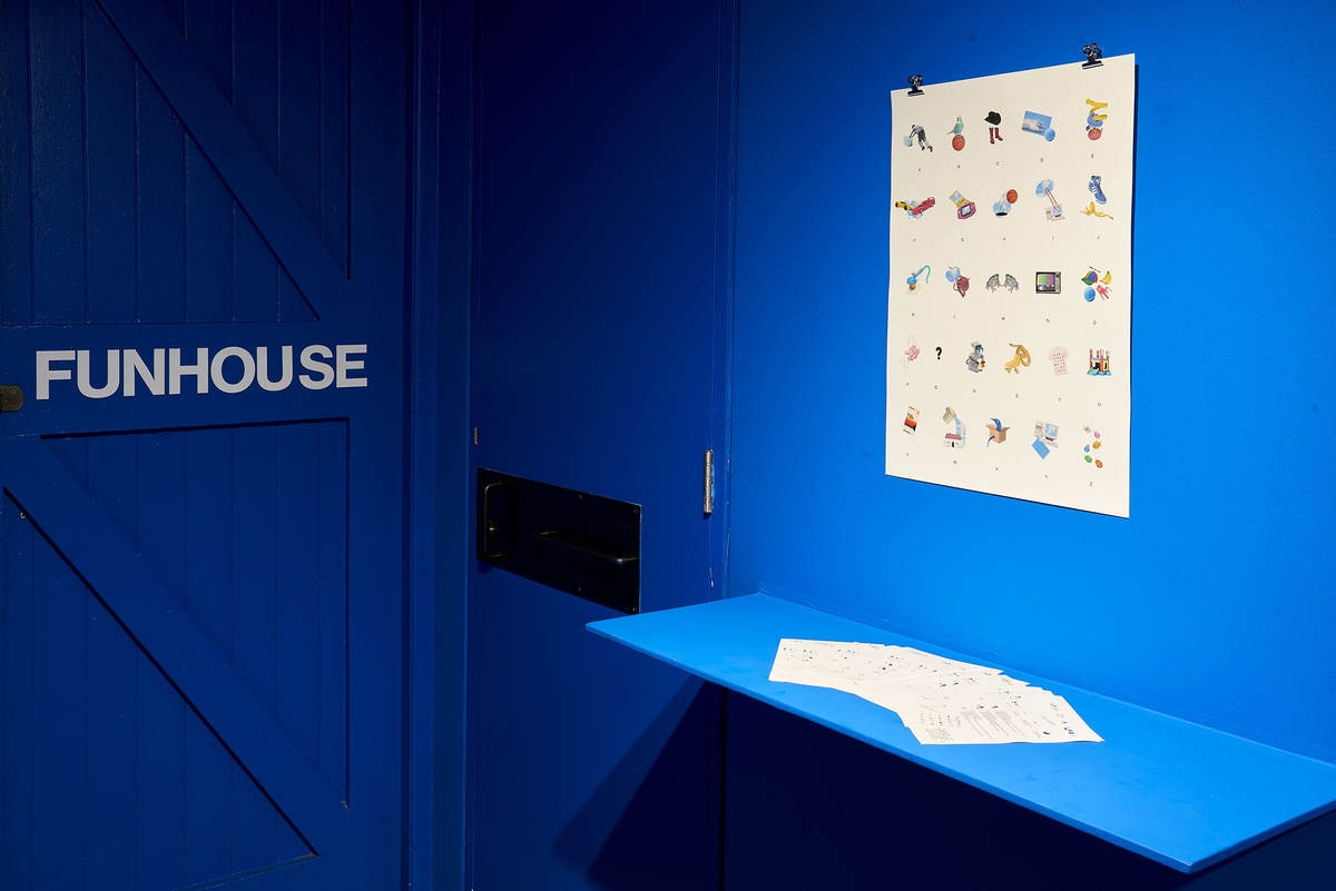 Installation photograph from the ‘Funhouse’ exhibition in A4’s Goods project space that features Luca Evans’ ‘Object Font’ project. A poster mounted on the blue wall features a grid of illustrations and characters from the latin alphabet. A blue wall-mounted shelf below it holds A4 size printouts with text and images.
