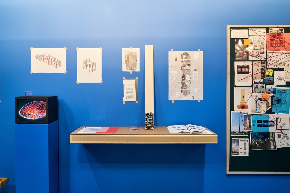 Installation photograph from the ‘mode(l)’ exhibition in A4’s Goods project space. On the left, Nkhensani Mkhari’s video ‘Soft Machine sample’ is displayed on a blue plinth. In the middle, a cardboard shelf features Thelma Ndebele’s ‘Groove Bienalle’ prints, Bonolo Kavula’s untitled paper and Shweshwe fabric sculpture and Gio Rech’s printed thesis ‘Homing: Delineating Liminal Constitutions’.
