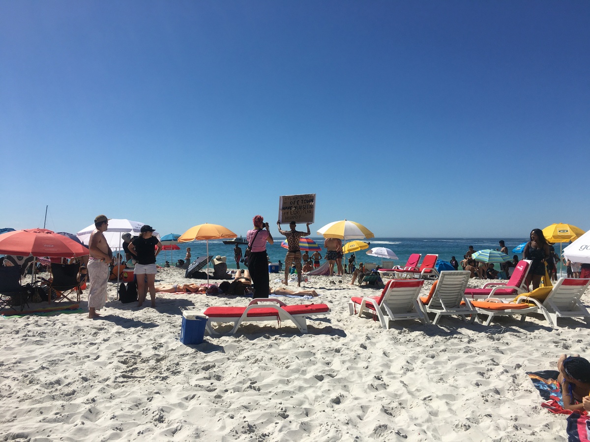 Event photograph from Kieron Jina’s ‘Catching Feelings, not Flights’ performance. The artist, their partially nude body covered in geometric designs, walks next to a Cape Town beach carrying a canvas that reads ‘It’s hunting season in Cape Town, have you seen my European husband?’ 
