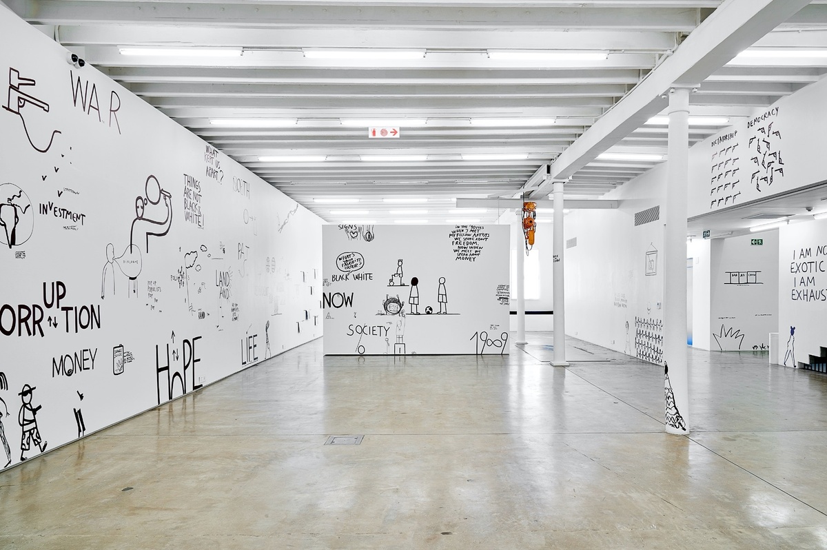 Installation photograph from Dan Perjovschi’s ‘The Black and White Cape Town Report’ exhibition in A4’s Gallery. On the left, the gallery is covered in drawings and phrases in black felt pen marker, including phrases like ‘war’, ‘hope’, ‘life’ and ‘corruption’. At the back, a moveable gallery wall is covered in drawings and phrases in black felt pen marker, including figures with soccer balls and phrases like ‘society’.
