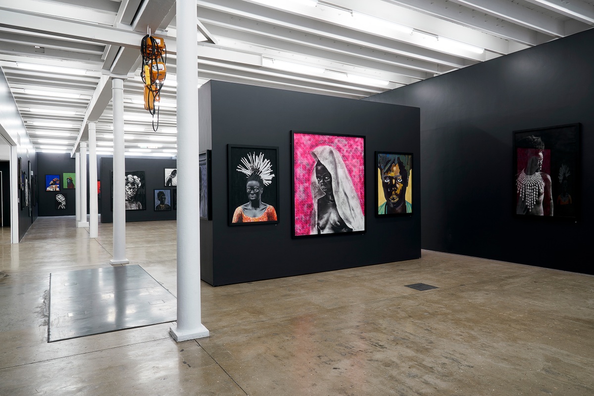 Installation photograph from the “Ikhono LaseNatali” exhibition in A4’s Gallery. In the middle, Major Ndlovu’s acrylic painting “Somnyama Ngonyama, ‘Ndivile II’,” Mlamuli Sboniso Mkwanazi’s charcoal drawing “Somnyama Ngonyama,” and Nomusa Mtshali’s acrylic painting “Somnyama Ngonyama, ‘Musa’” are mounted on a black moveable gallery wall. On the right, Nhlankanipho Mkhize’s acrylic painting “Somnyama Ngonyama” is mounted on the black gallery wall.
