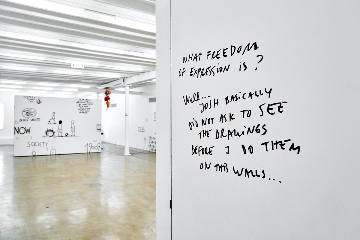 Installation photograph from Dan Perjovschi’s ‘The Black and White Cape Town Report’ exhibition in A4’s Gallery. On the right, a white wall features writing in black felt pen marker, reading “What freedom of expression is? Well … Josh basically did not ask to see the drawings before I do them on this walls …”

