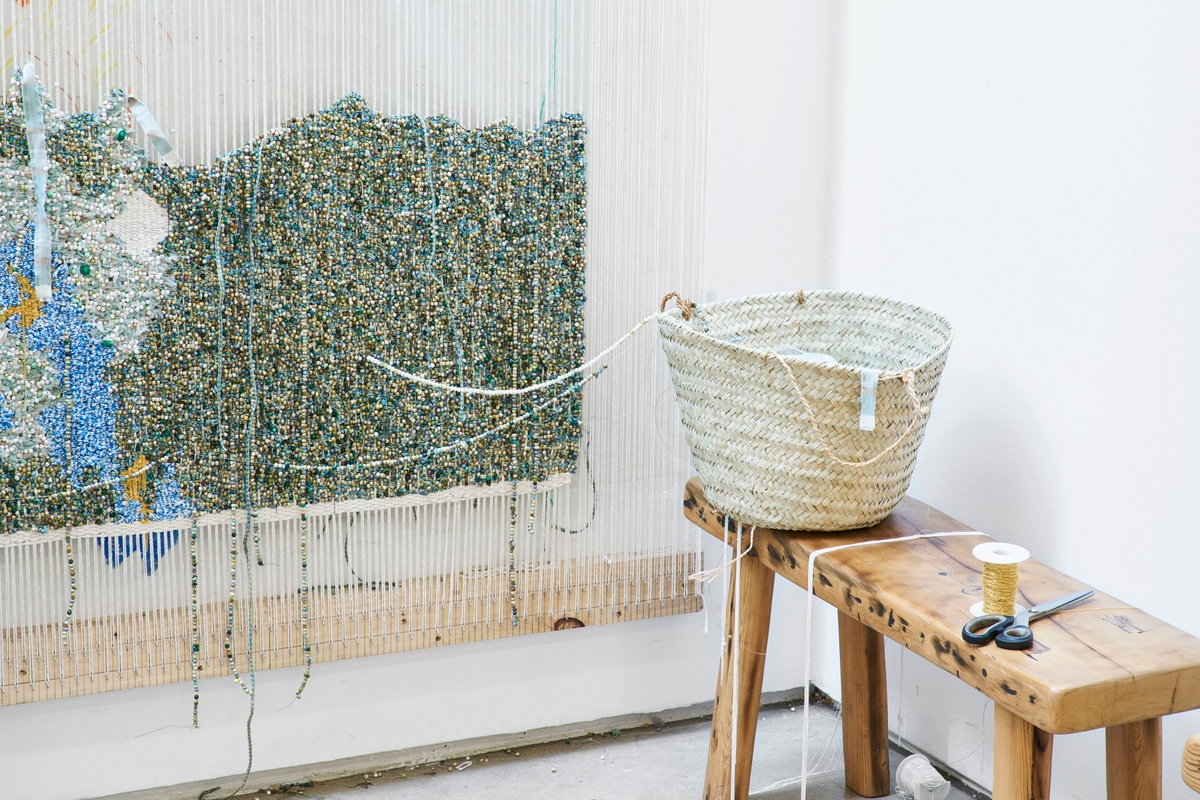 Process photograph from ‘Open Production’, Igshaan Adams’ hybrid studio/exhibition in A4’s Gallery. On the right, a wooden bench with woven basket and scissors. On the left, a mixed media tapestry in the process of being woven is mounted on the wall.
