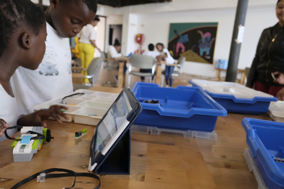 Event photograph from the Lego Robotics workshop by ORT SA CAPE on A4’s top floor. A table covered with plastic containers, lego electronic components and a digital tablet is surrounded by children.
