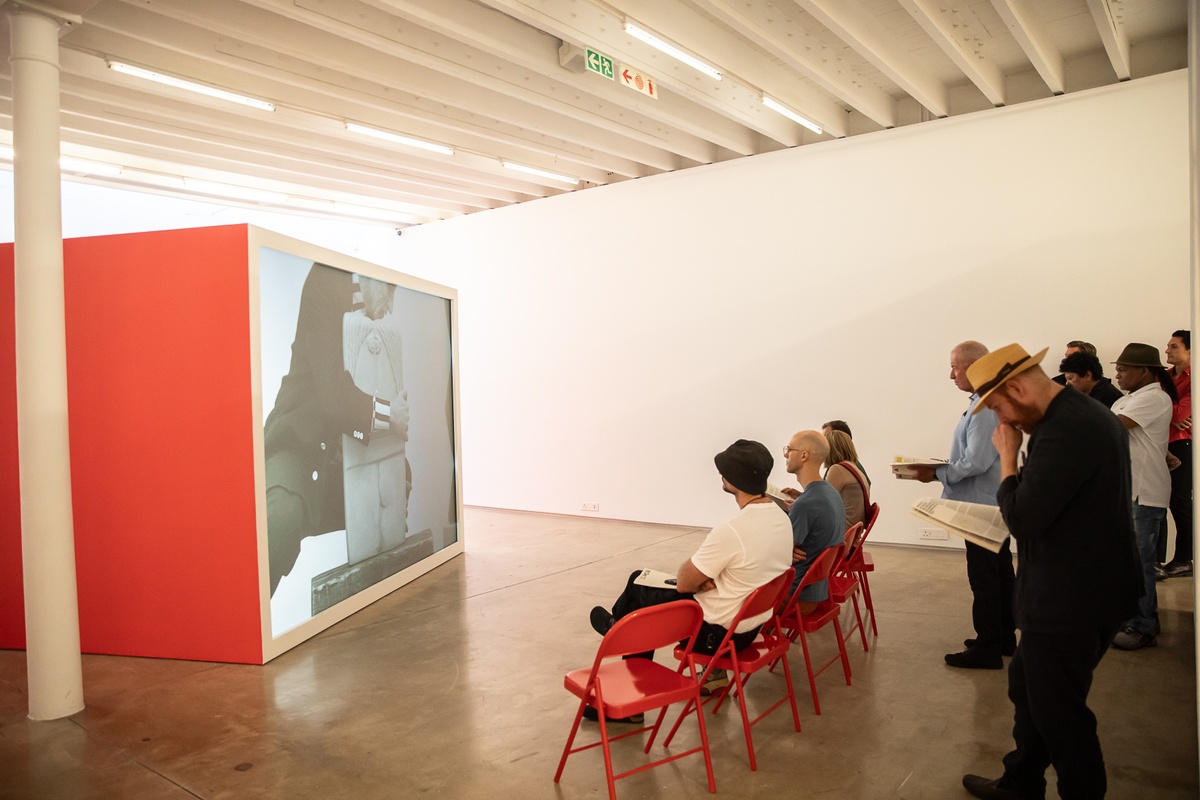 Event photograph from the preview of the 'A Little After This' exhibition in A4 Arts Foundation that shows Alex Da Corte's video installation 'ROY G BIV'. On the left, a large red wooden box with a back-projected screen plays Da Corte's video. On the right, 7 red powder-coated viewing chairs are arranged in an arch, with attendees seated on them and standing behind them.
