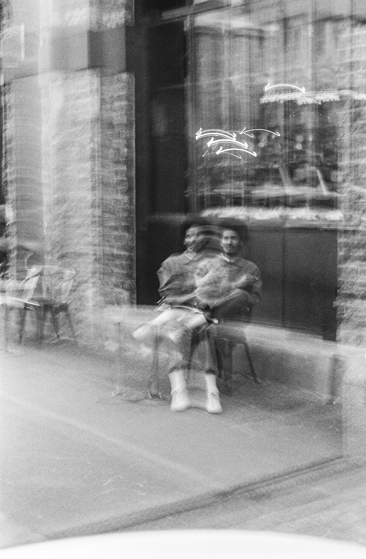 Monochrome event photograph from ‘Contact’, a collaboration between A4’s proto~ museum shop and Oath Magazine, taken by a participant. A motion-blurred photograph of a person sitting outdoors on a chair in front of a series of windows.
