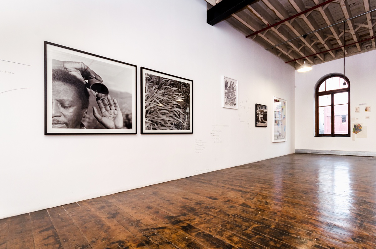 Installation photograph from the ‘Between Two or More Worlds’ exhibition at the 2016/2017 A4 Office that shows several wall mounted artworks interspersed with charcoal drawings and writing. On the left, George Hallet’s photographic diptych ‘Peter Clarke’s Tongue’, Thabiso Sekgala’s photograph ‘The Terrace Hotel, Hulawayo’ and Moshekwa Langa’s mixed media work ‘Untitled (Objects in a Mirror)’ line the white wall.
