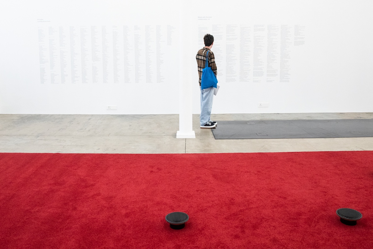 Event photograph from the opening of The Future Is Behind Us exhibition in A4’s gallery. At the front, James Webb’s sound installation ‘Prayer’ is visible, with red carpeting and two upturned speakers. At the back, an individual reading the vinyl wall text credits for ‘Prayer’.
