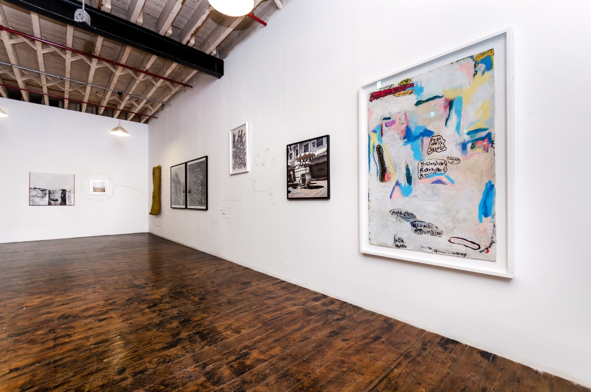 Installation photograph from the ‘Between Two or More Worlds’ exhibition at the 2016/2017 A4 Office that shows several wall mounted artworks interspersed with charcoal drawings and writing. On the left, Gerda Scheepers’ acrylic painting ‘Pondoland pocket’ and Thabiso Sekgala’s photograph ‘Semotlhase, former Bophuthatswana’ line the white wall. On the right, Bronwyn Katz’s bed spring and mattress hanging sculpture ‘Droom boek’, George Hallet’s photographic diptych ‘Peter Clarke’s Tongue’, Thabiso Sekgala’s photograph ‘The Terrace Hotel, Hulawayo’ and Moshekwa Langa’s mixed media work ‘Untitled (Objects in a Mirror)’ line the white wall.
