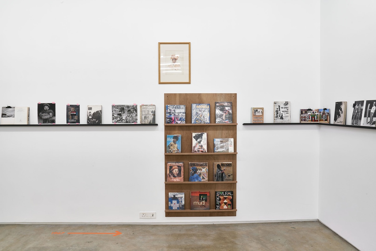 Installation photograph from the Photo Book! Photo-Book! Photobook! exhibition in A4’s Gallery. In the middle, George Pemba’s watercolour painting ‘Basotho Woman with Headdress’ hangs above a display shelf in an area dedicated to photobooks from the years 1967 to 1994.
