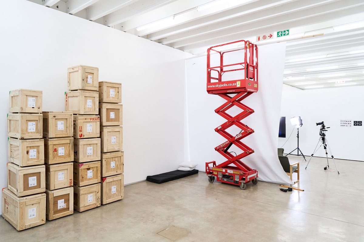 Process photograph from the 2nd rendition of ‘Parallel Play’ in A4’s Gallery. On the left, wooden crates are stacked against the white wall. On the right, an aerial work platform and makeshift photography studio.
