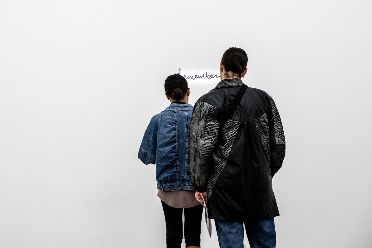 Event photograph from opening of the Customs exhibition in A4’s Gallery that shows two attendees standing in front of Steve McQueen’s neon tube and acrylic paint installation ‘Remember Me’ mounted on a white gallery wall.
