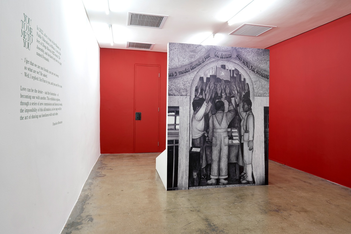 Installation photograph of the You to Me, Me to You exhibition. On the left, the exhibition title and blurb are pasted onto the wall in green vinyl. On the right, Tina Modotti’s photograph ‘La protesta. Corrido de la Revolución.’ is reproduced as wallpaper on a freestanding wall.
