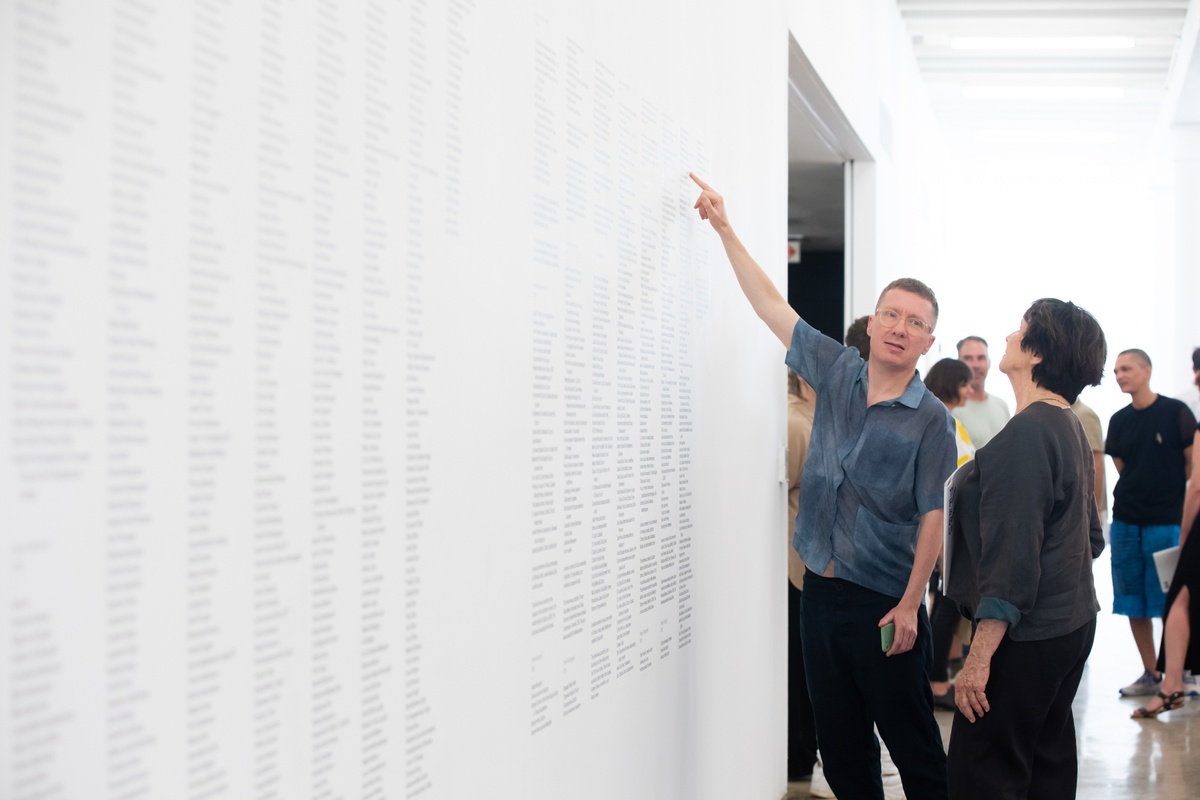 Event photograph from the opening of The Future Is Behind Us exhibition in A4’s gallery. Artist James Webb points out a detail on the vinyl wall credits for his installation ‘Prayer’ to Sue Williamson.
