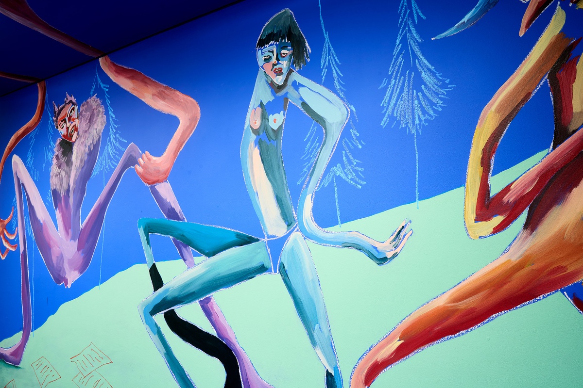 Installation photograph of Dominique Cheminais’ residency in A4’s Goods project space. A painted mural with humanoid figures lines the blue walls.
