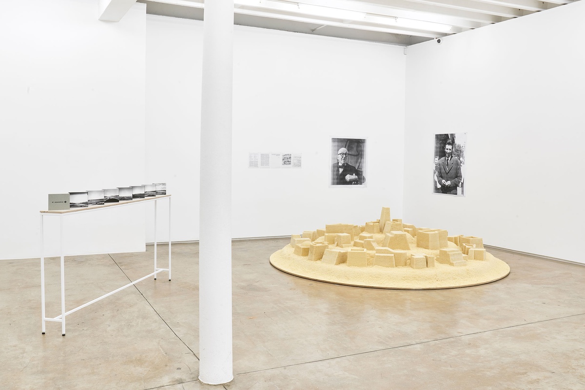 Installation photograph from the The Future Is Behind Us exhibition in A4’s Gallery. On the left, Jo Ractliffe’s open-spine accordion book ‘N1: every 100 kilometers’ sits on a thin rectangular table. On the right, Kader Attia’s couscous sculpture ‘Untitled (Ghardaïa)’ sits on the gallery floor.
