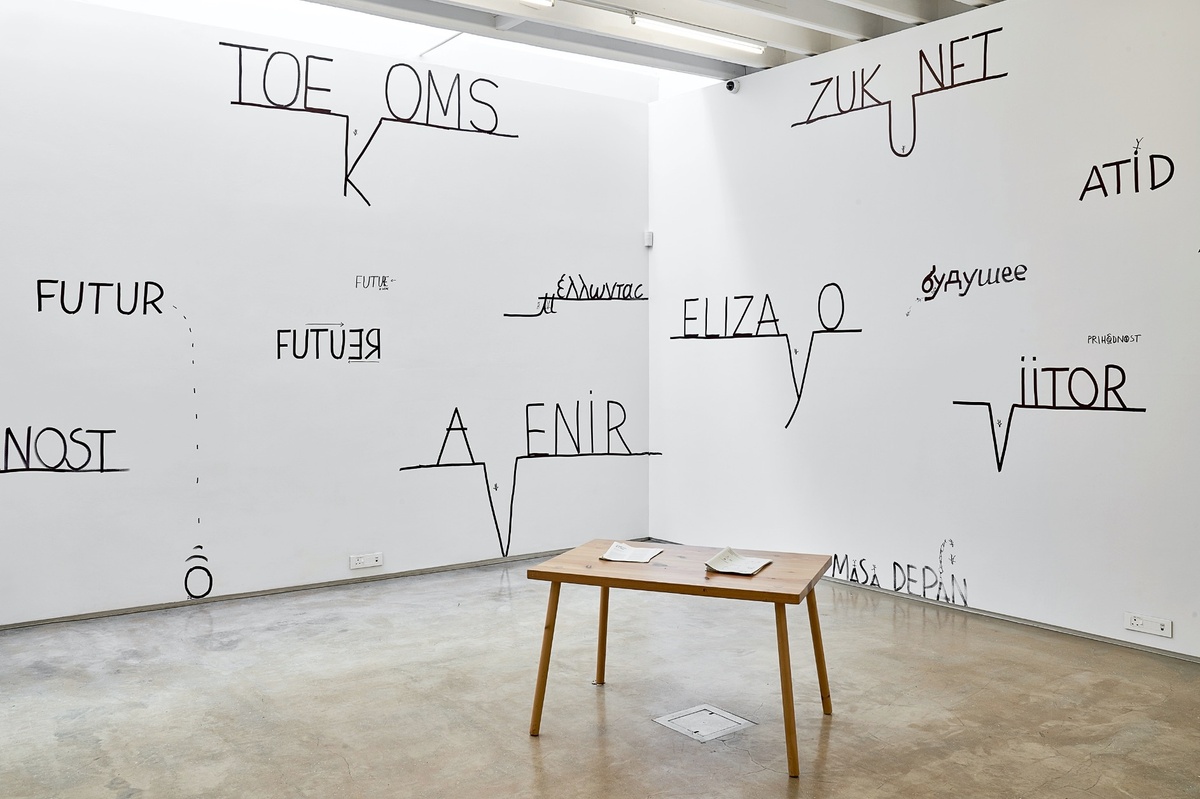 Installation photograph from Dan Perjovschi’s ‘The Black and White Cape Town Report’ exhibition in A4’s Gallery. At the front, a wooden table with booklets. At the back, the wall holds several translations of the word ‘future’ on top of lines, where one character is dropped into the line so as to resemble a geographic feature.
