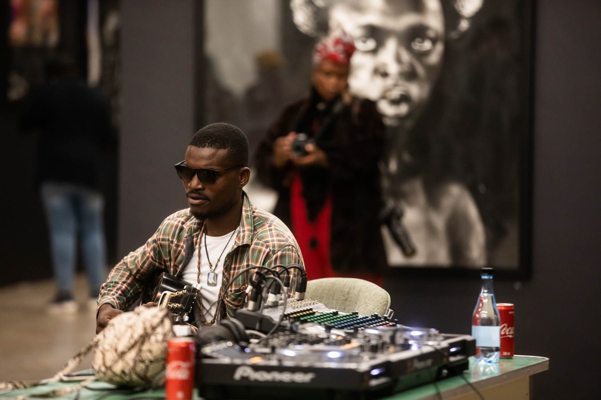 Event photograph from ‘Finissage: Ikhono LaseNatali’ in A4’s Gallery. At the front, a guitarist sits behind a table laden with audio equipment. At the back, a photographer stands in front of Mlamuli Sboniso Mkwanazi’s wall-mounted charcoal on paper drawing ‘Somnyama Ngonyama’.
