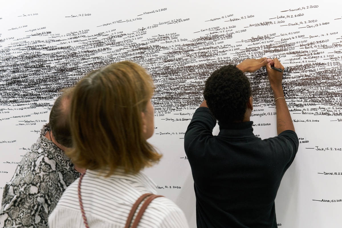 Event photograph of Roman Ondák’s performance piece ‘Measuring the Universe’ at A4’s booth at the 2020 Cape Town Art Fair. At the back, a white wall is covered with overlapping marks that indicate participant’s height, names and the date of their participation in black felt pen. At the front, A4’s Obakeng Motsepe ads information to the wall, with two fair attendees behind him.
