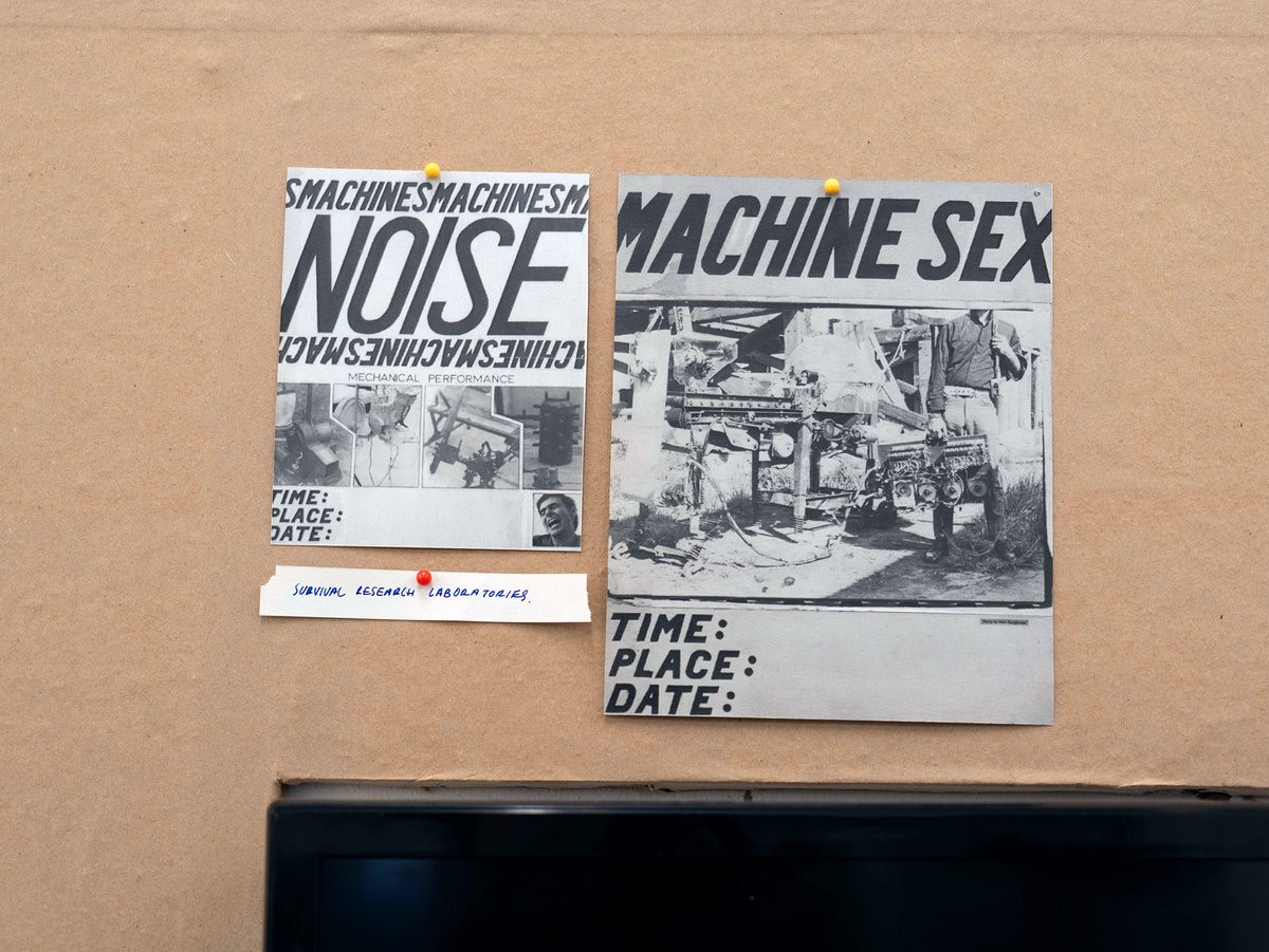Installation photograph from Mitchell Gilbert Messina's residency in A4 Art Foundation shows a wall-mounted strip of cardboard hosting pinned research notes. Posters for two events hosted by Survival Research Laboratories, an organisation of artists and creative technicians founded in 1978 by Mark Pauline.
