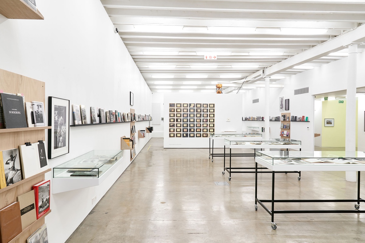 Installation photograph from the Photo Book! Photo-Book! Photobook! exhibition in A4’s Gallery. On the left, shelving units with printed matter from the years 1967 to 1994 line the gallery wall, along with David Goldblatt’s photograph ‘Climbing into kibbles at the shaft-head, the shift gets ready to go down. President Steyn No.4 shaft, Welkom, Orange Free State. 1969’. On the right, three freestanding glass display cases with printed matter. At the back, Sue Williamson’s print series ‘For Thirty Years Next to His Heart’ is mounted on a white moveable gallery wall in a grid.
