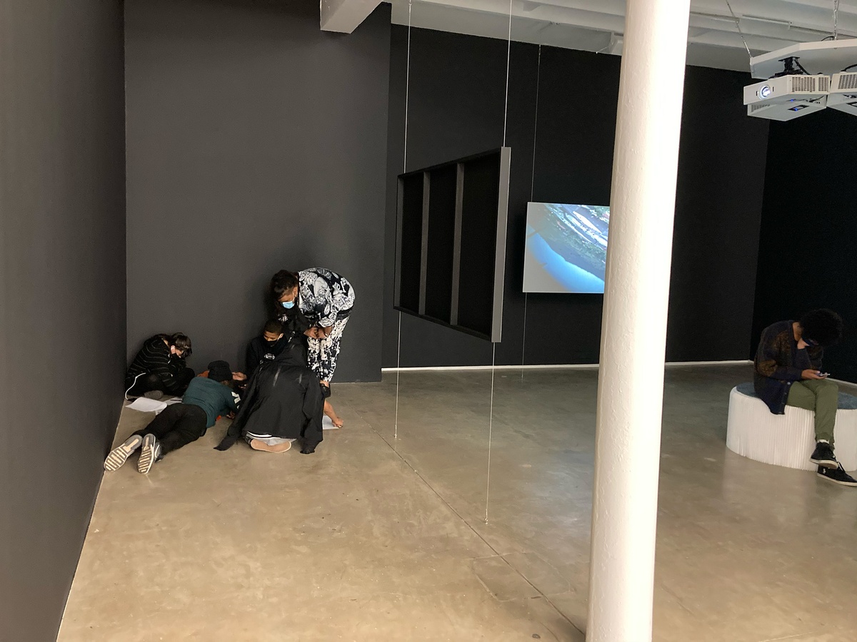 Event photograph from the ‘Lalela x A4' exchange during the ‘Tell It to the Mountains’ exhibition in A4’s Gallery. On the left, Lalela students huddle together in a corner of the gallery. On the right, Mikhael Subotzky’s video installation ‘Moses and Griffiths’.
