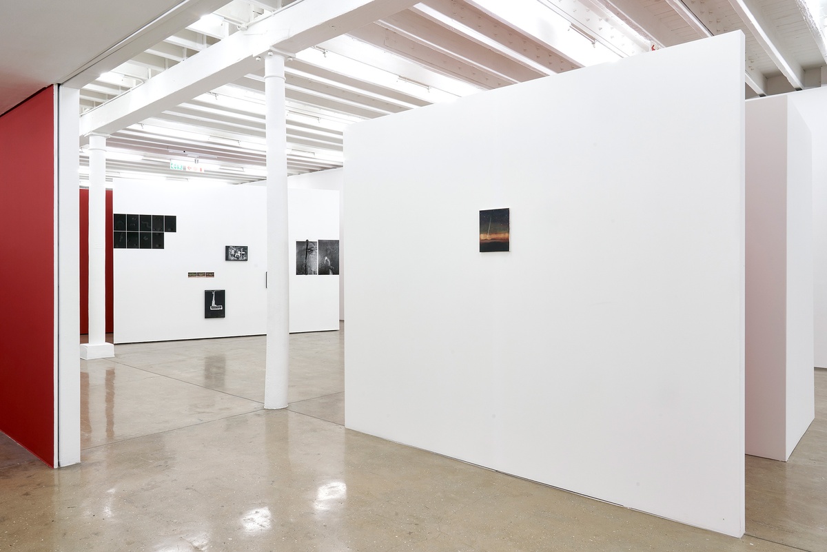 Installation photograph of the You to Me, Me to You exhibition. On the right, Dexter Dalwood’s oil painting ‘Francisco Fernando’ is mounted on a white freestanding wall. On the left, Thembinkosi Hlatshwayo’s numerous photographic works are arranged on a white moveable gallery wall.
