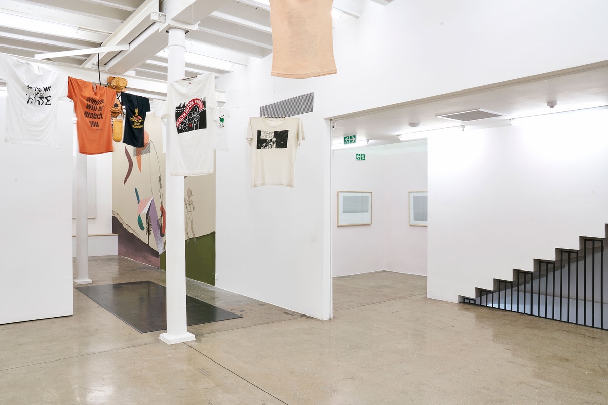 Installation photograph of the Common exhibition. Above on the left, T-shirts with slogans from the GALA Queer Archive and SAHA are suspended on criss-crossing lines. In the middle towards the back are framed Fabian Saptouw prints from their ISBN University series with layered ISBN and ISSN numbers hung on a pale pink wall.
