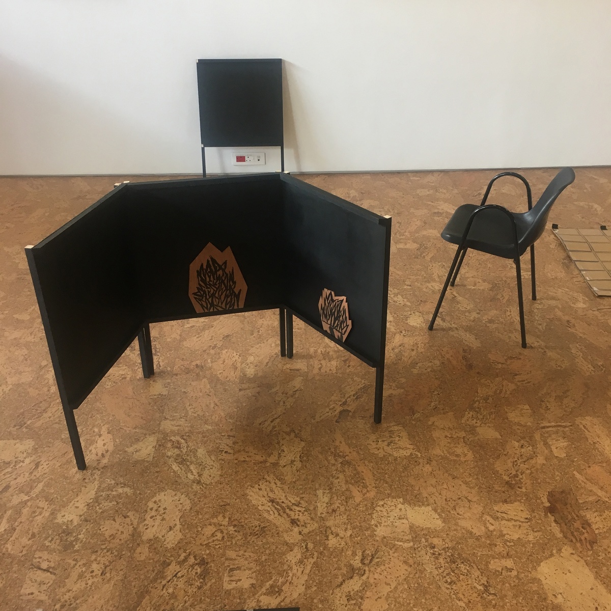 Process photograph from Jonah Sack’s residency on A4’s top floor. A small sculptural object on the floor consists of three small black panels on stands, interlinked to resemble a hearth. The middle and righthand side panels hold small drawings that resemble flames.
