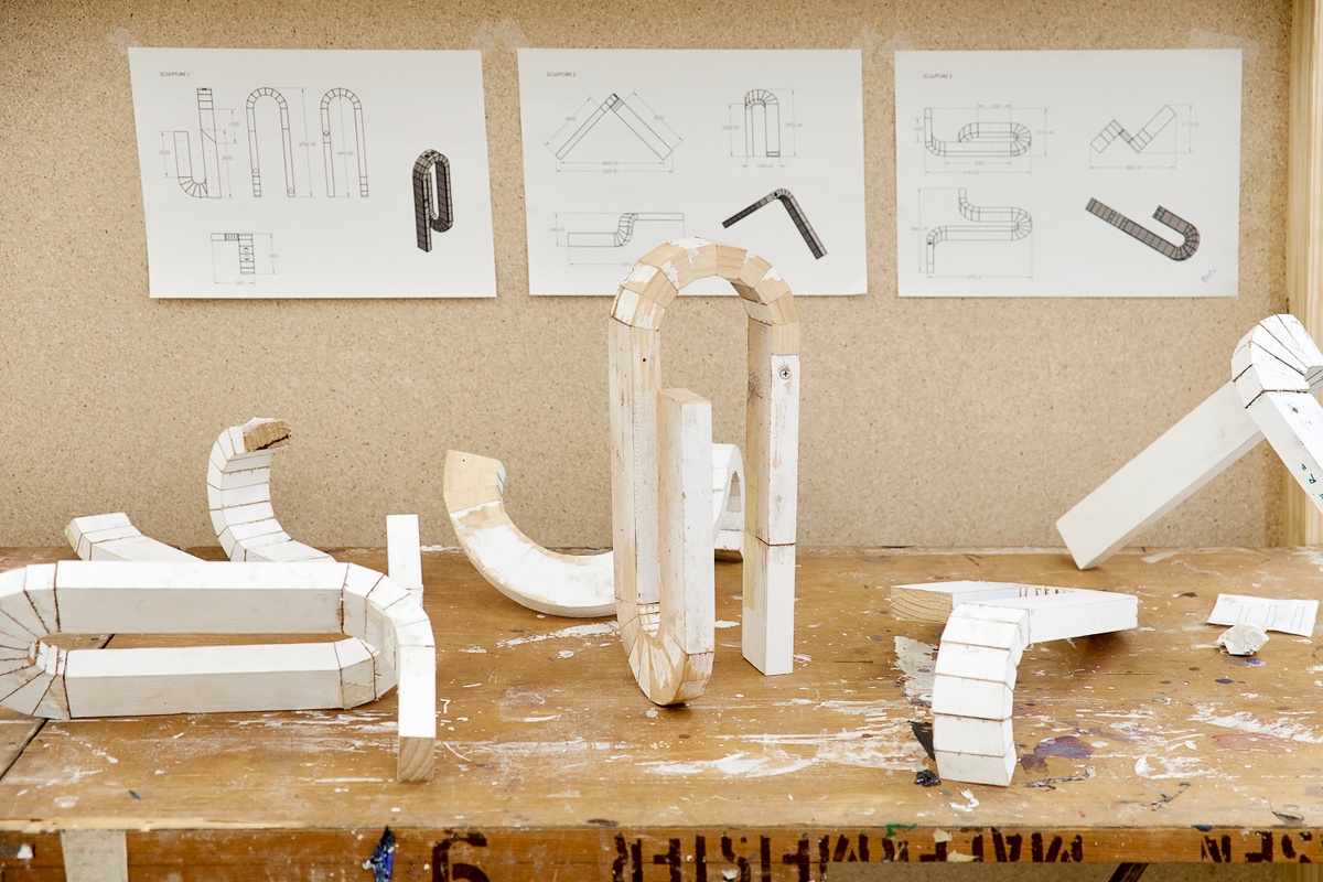 Installation photograph from the 2018 rendition of ‘Parallel Play’ in A4’s Gallery. At the front, white painted wooden prototypes from Kyle Morland’s sculptural practice sit on a wooden table. At the back, diagrams for the prototypes are taped to a wooden stand.
