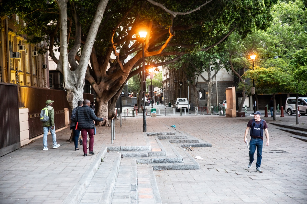 Process photograph from the 2019 rendition of Christian Nerf’s performance piece ‘Polite Force’. On the left, a participant wearing ‘Polite Force’ riot gear walks along a paved walkway. On the right, members of the public walk in the opposite direction.
