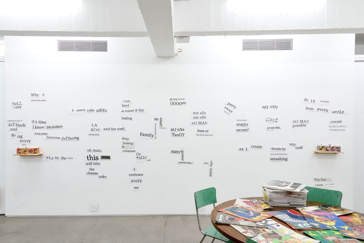 Installation photograph from the Papertrails exhibition in A4’s Reading Room. On the right, a round table strewn with printed matter. At the back, printed word and sentence snippets are pasted onto the wall.

