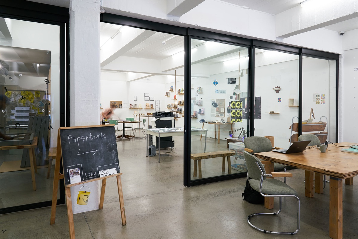 Installation photograph from the Papertrails exhibition in A4’s Reading Room. On the left, a small blackboard with the exhibition title in chalk. On the right, wooden tables with seating. In the middle, the Papertrails working studio.
