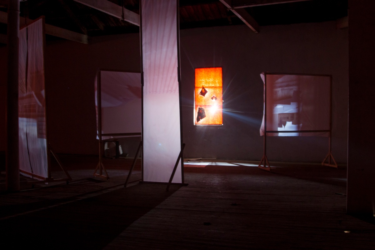 Installation photograph from George Mahashe’s ‘Camera Obscura #3’ exhibition on A4’s top floor. The darkened space features freestanding surfaces with a projected images from A4’s exterior facilitated by Mahashe’s pinhole camera. In the middle, a covered window lets in a ray of light.
