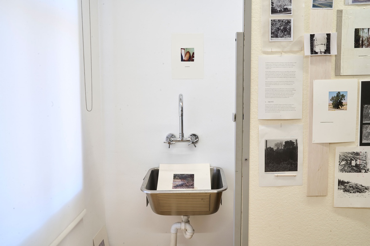 Installation photograph of Michael Tymbios’ studio during his residency in A4 Arts Foundation. On the left, a piece of backing board with a photographic print sits in a metal sink. On the right, a freestanding pinboard features multiple text printouts and photographic prints.
