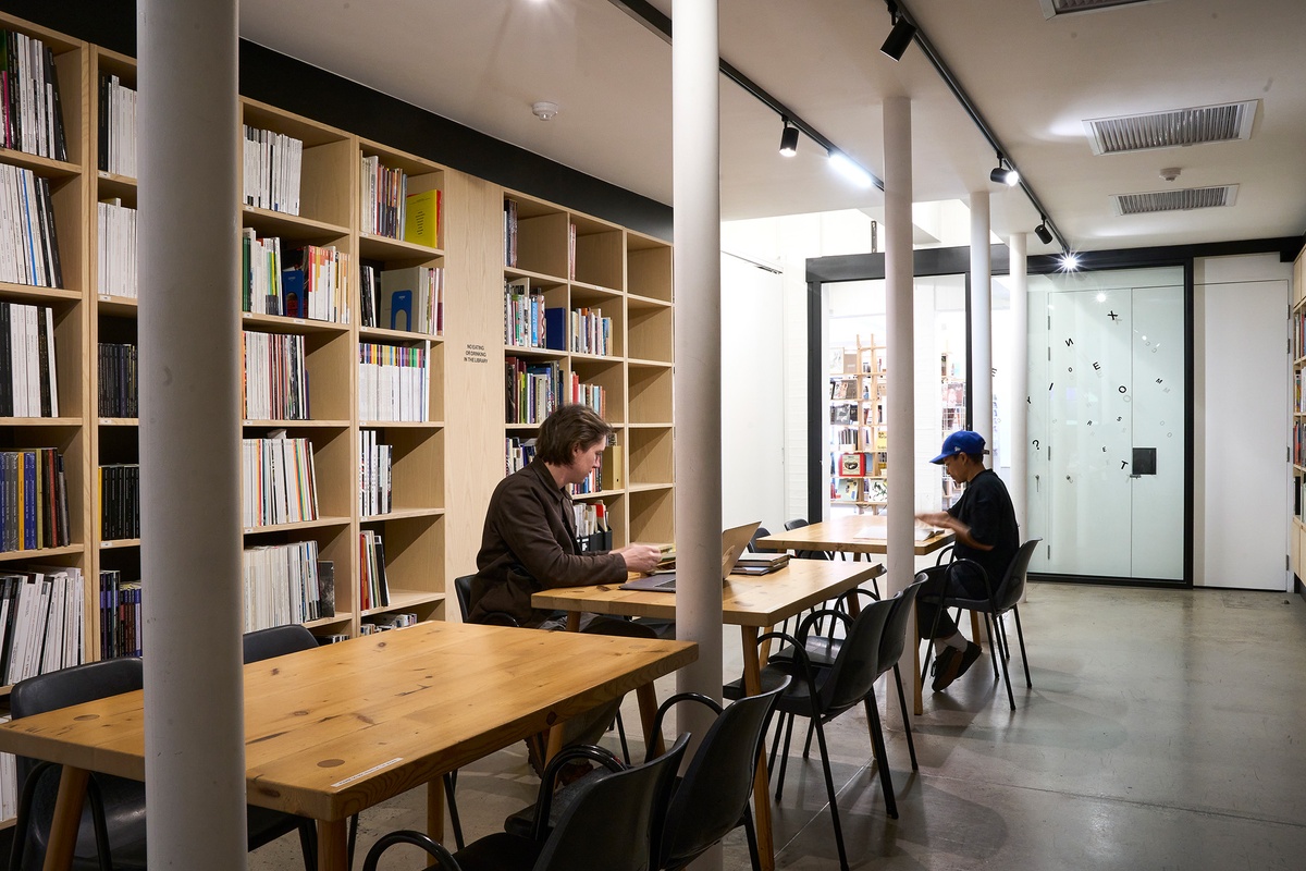 A photograph of the A4 Library. In the middle, a row of rectangular wooden tables with chairs. On the left, a row of book shelves. At the back, a sliding glass door.
