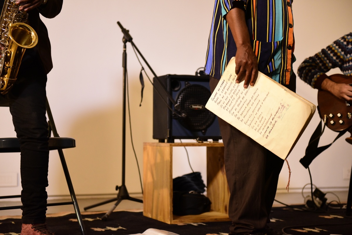 Event photograph from ‘Between Space & Time’ musical performance on A4’s ground floor. At the front, one of the participating musicians holds a notebook with lyrics written on it. At the back, an electronic amplifier sits on a wooden box.
