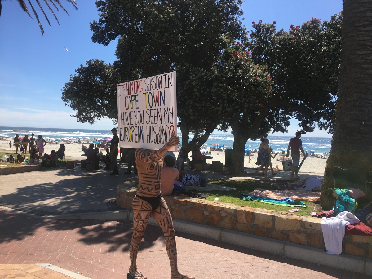 Event photograph from Kieron Jina’s ‘Catching Feelings, not Flights’ performance. The artist, their partially nude body covered in geometric designs, walks next to a Cape Town beach carrying a canvas that reads ‘It’s hunting season in Cape Town, have you seen my European husband?’ 
